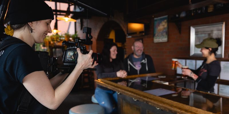 Danielle Hazelton filming a bartender serving drinks to a smiling couple at a cozy bar.